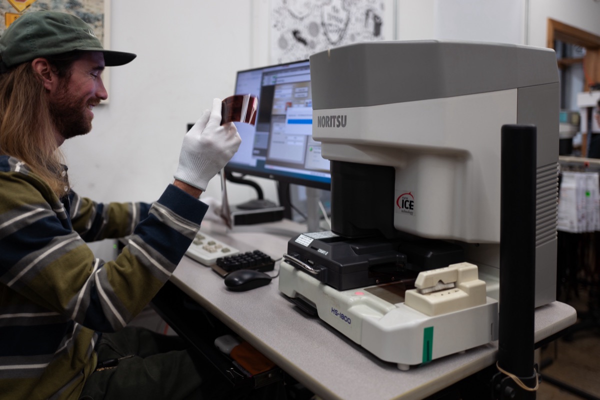 A Darkroom employee in white gloves checks a strip of negatives beside a Noritsu HS-1800 film scanner